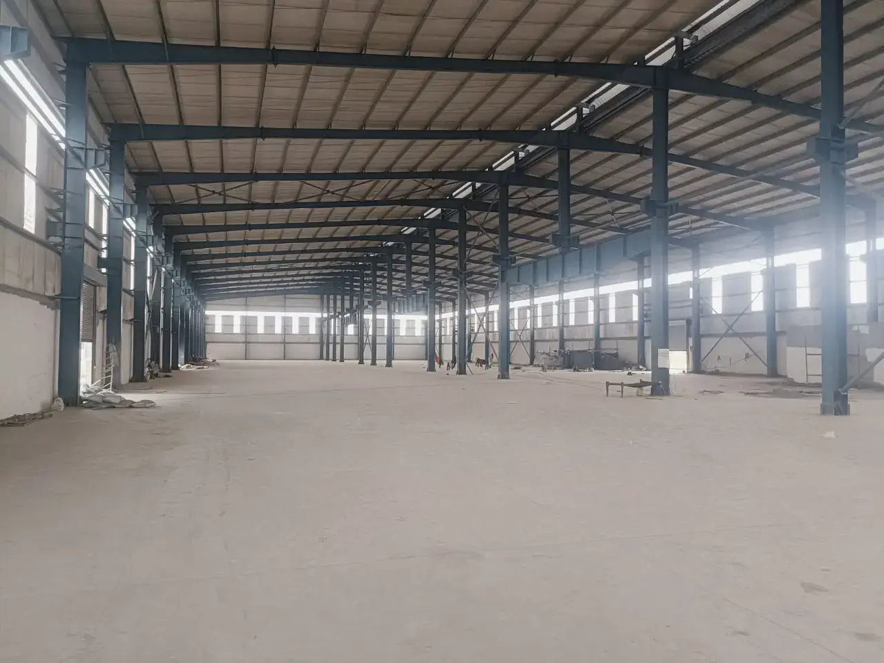 Empty warehouse interior with concrete floor, steel columns and roof trusses, side windows, and scattered debris.