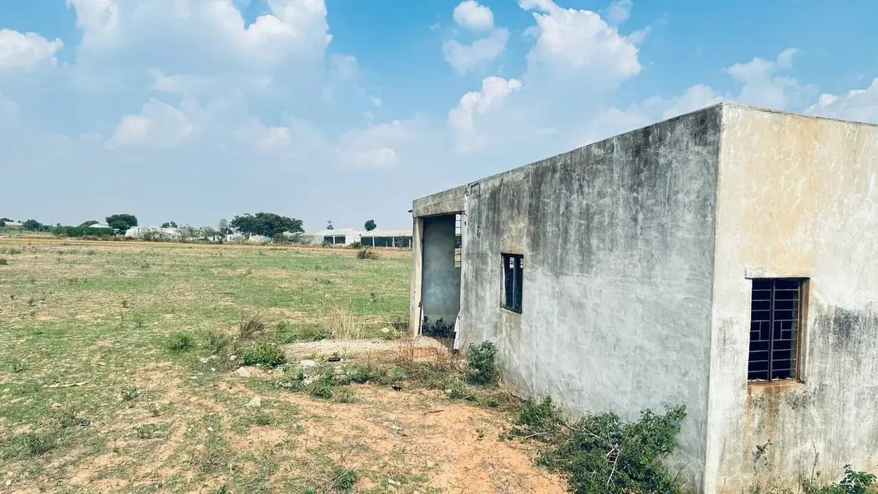 Exterior view of a small weathered concrete building beside an open grassy dirt field under a cloudy sky.