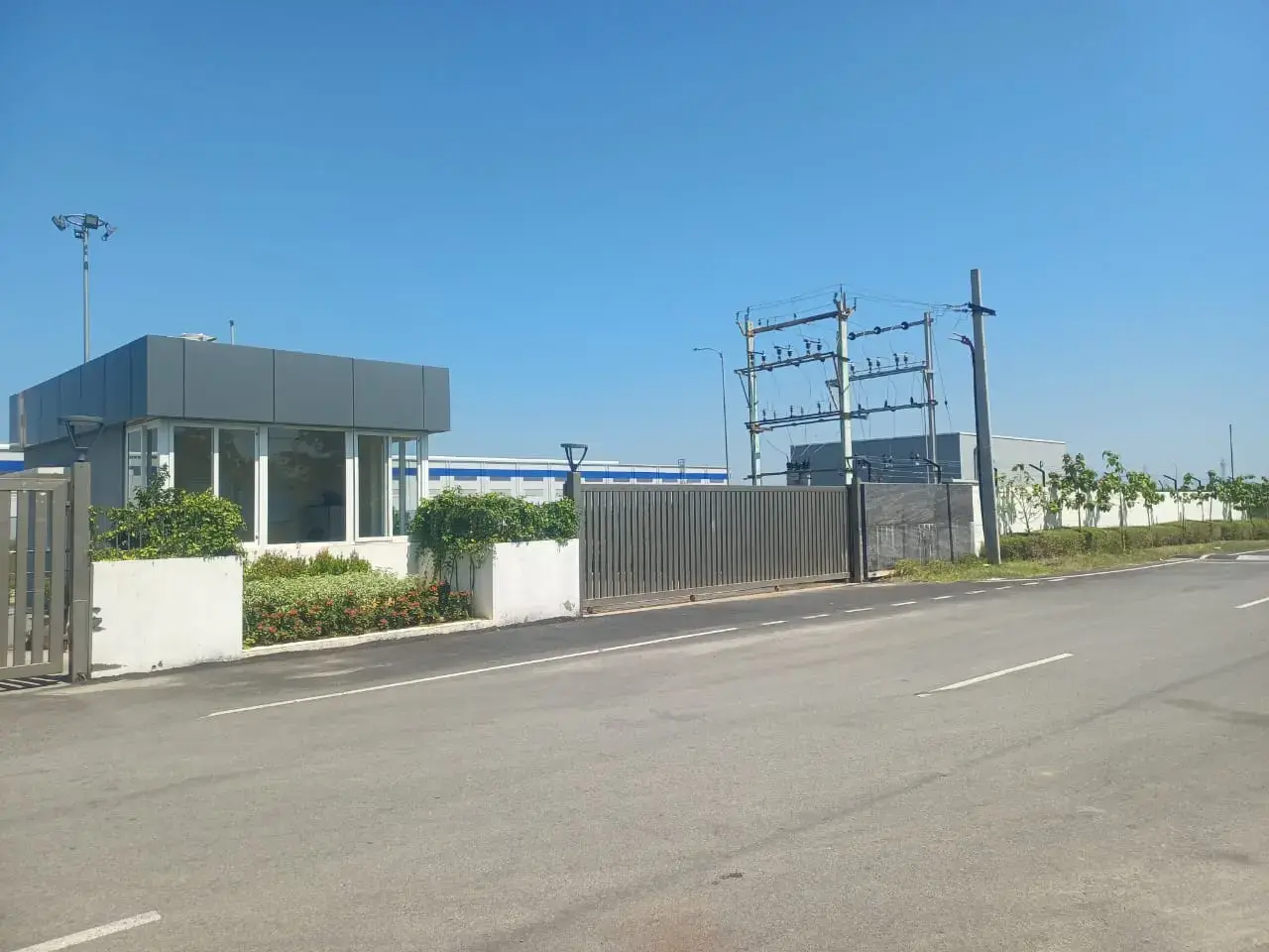 Paved road outside a gated industrial facility with a guard building, landscaping, boundary fence, and electrical equipment.
