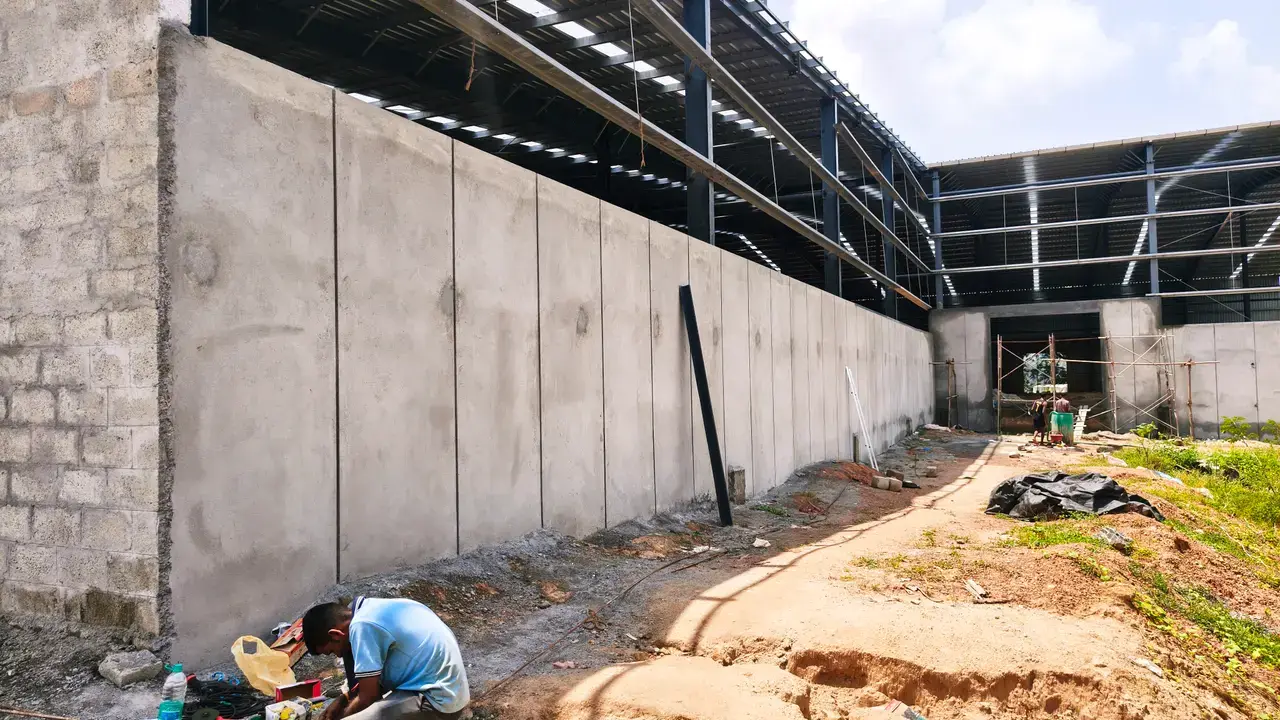 Exterior construction area beside a concrete wall and open steel-roofed structure, with dirt path, debris and a worker.