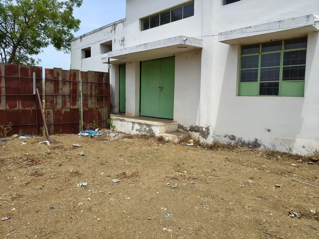 Exterior view of a white building with green doors and windows, a dirt yard, metal gate, and scattered debris.