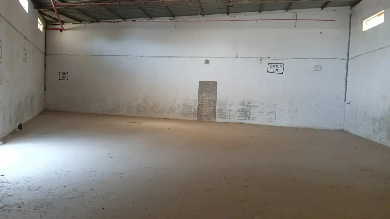 Empty warehouse interior with concrete floor, white walls, corrugated metal roof, small high windows, and a central doorway.