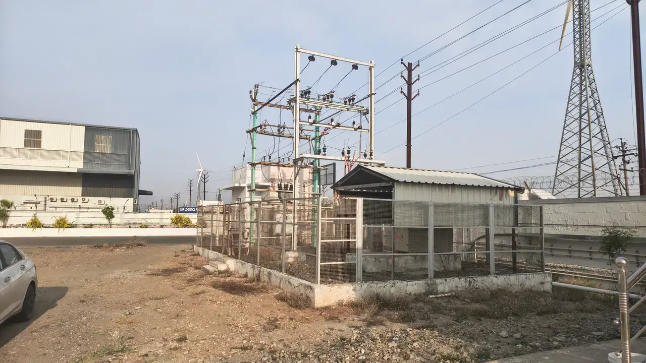 Gravel yard beside a fenced electrical substation, with utility poles, transmission tower, boundary wall and industrial sheds.