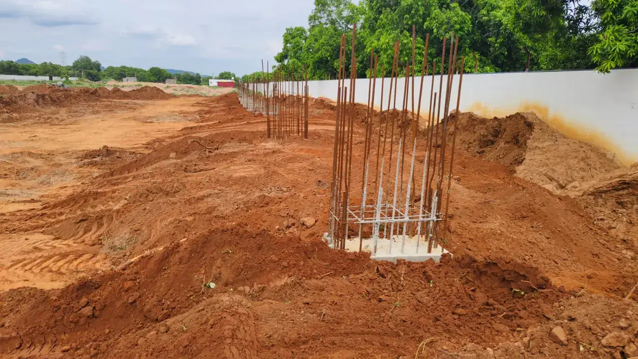 Open construction site with excavated earth, exposed reinforced concrete columns, a boundary wall and trees.
