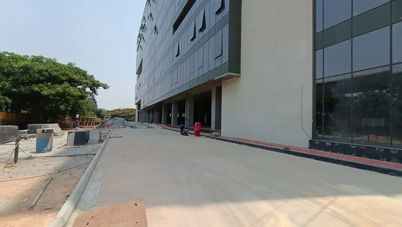 Paved driveway alongside a large building facade with glass windows, columns, construction materials, and trees.