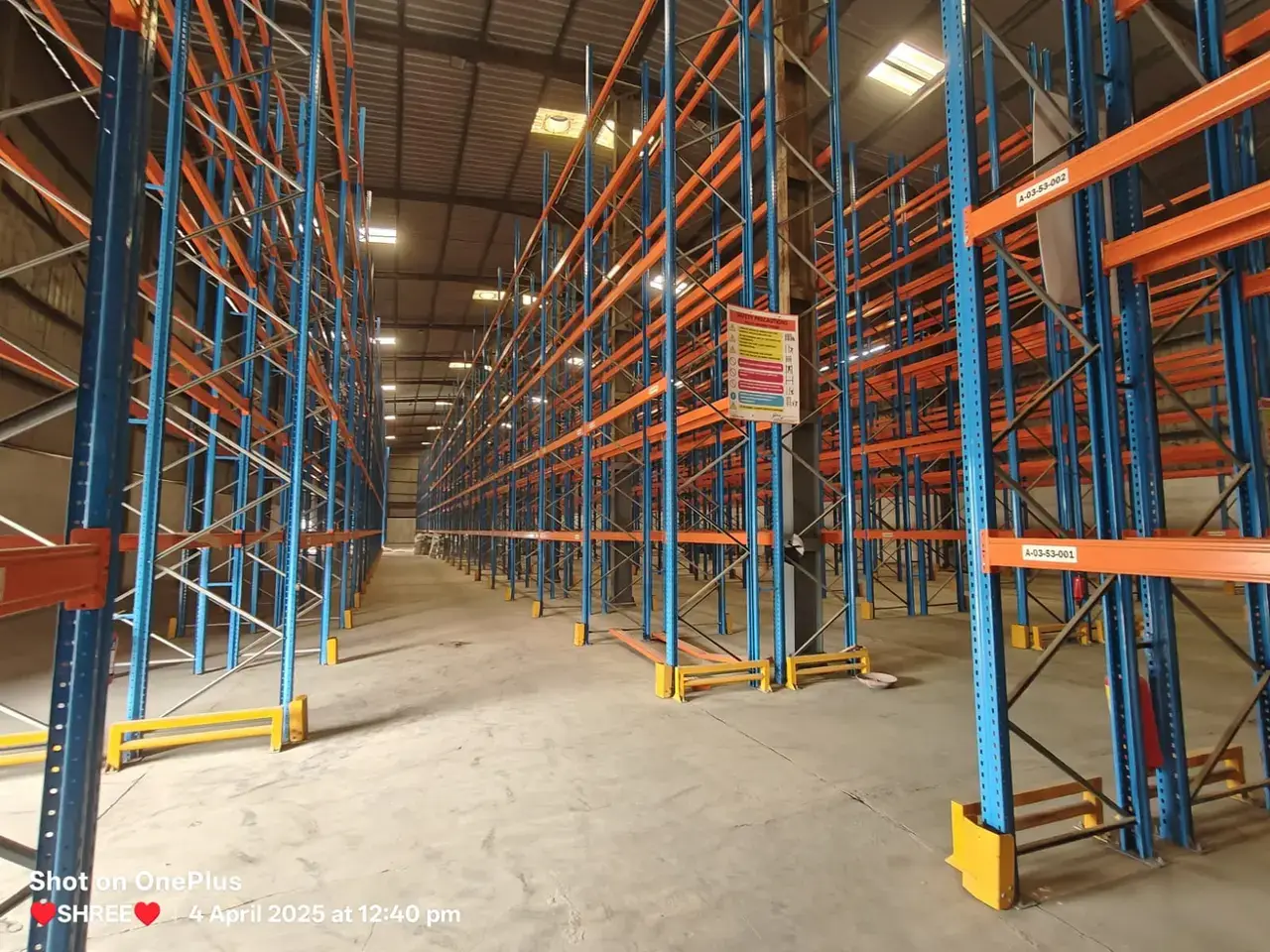 Warehouse interior with tall blue and orange pallet racking, concrete floor, roof lights, and empty storage aisles.
