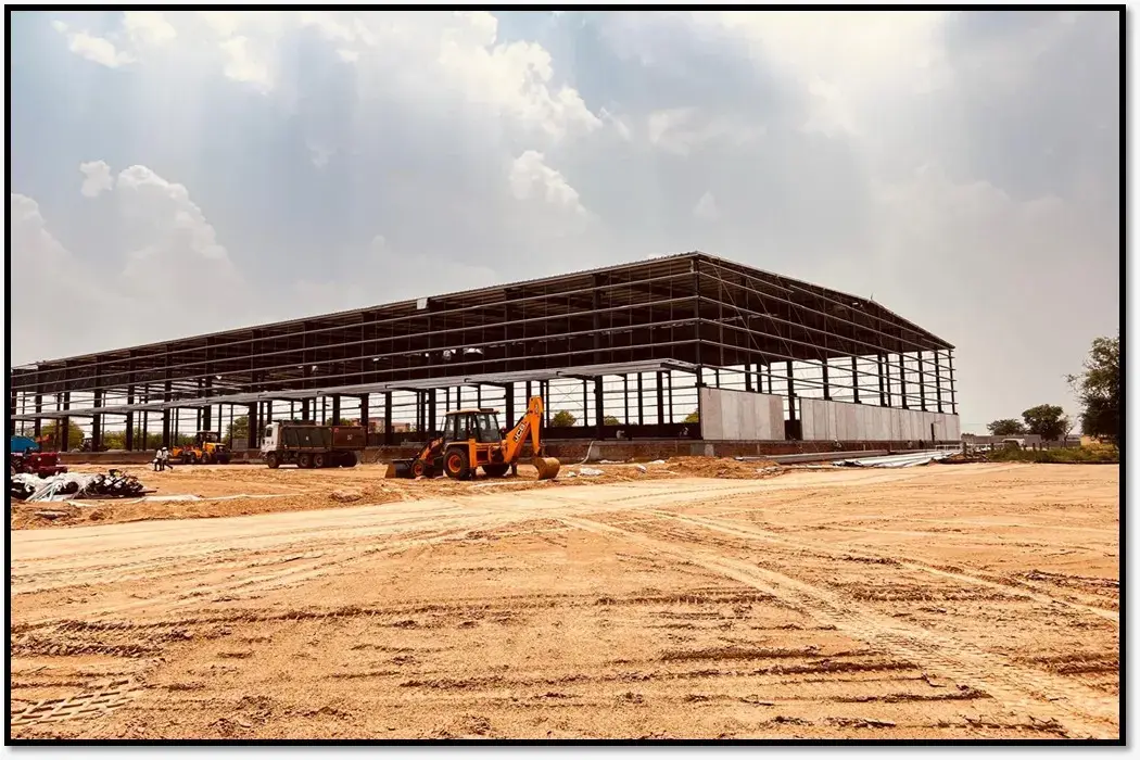 Open dirt yard facing a large steel-framed warehouse under construction, with construction equipment and cloudy sky.