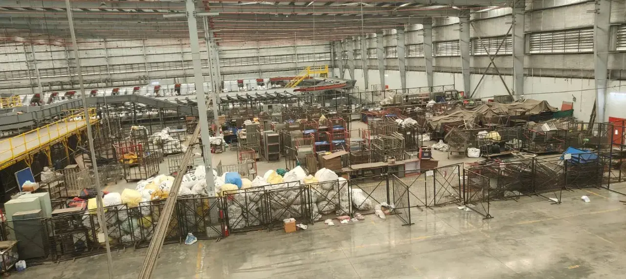 Large industrial shed interior with metal roof, columns, racks, machinery, stored materials and a concrete floor.