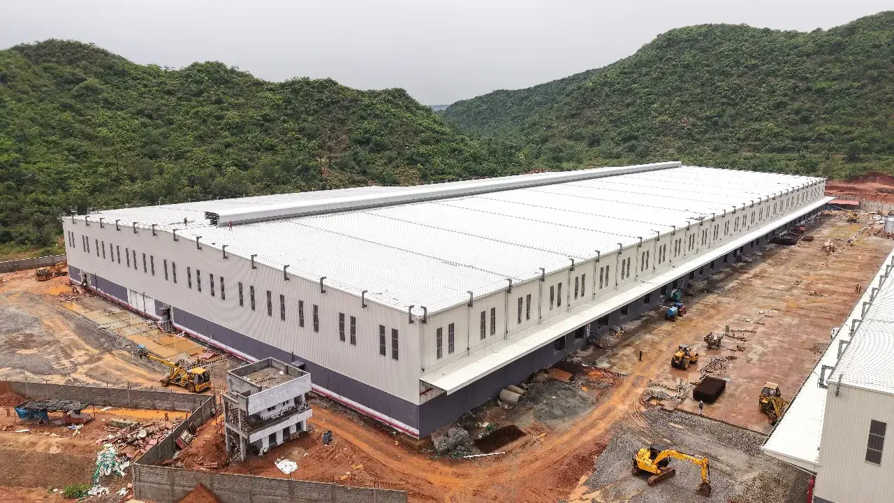 Aerial view of a large white industrial warehouse under construction, surrounded by dirt yards, machinery, and green hills.