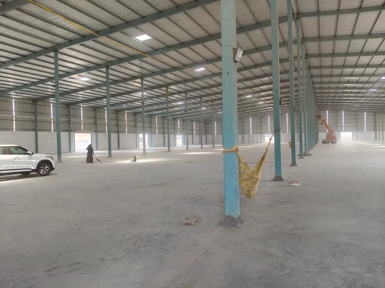Large empty warehouse interior with concrete floor, metal roof trusses, rows of blue columns, hanging fabric, and a parked car.
