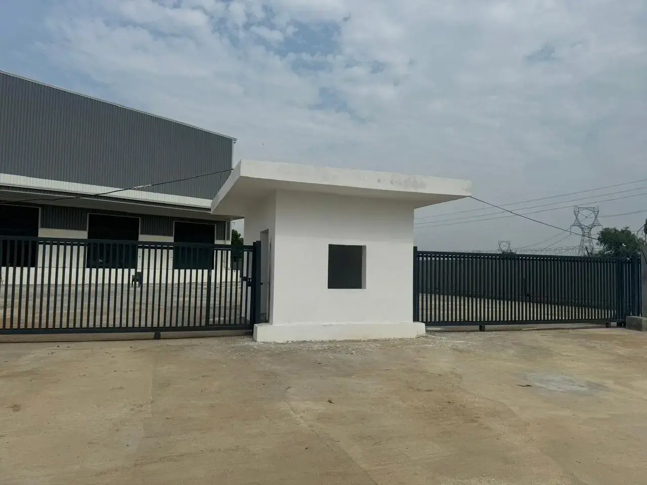 Warehouse entrance with black metal gates, a small white guardhouse, paved yard, and overhead power lines.